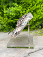 A piece of jewelry—possibly a necklace or bracelet—displayed on a clear stand. The jewelry appears to be made of a shiny material, possibly silver or gold, and has a unique, irregular shape. The background is blurred, but it appears to be a green plant or foliage, which contrasts with the metallic appearance of the jewelry.