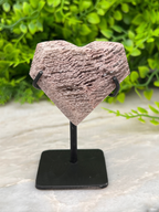 A heart-shaped rock sculpture is displayed on a black stand, set against a lush green background.