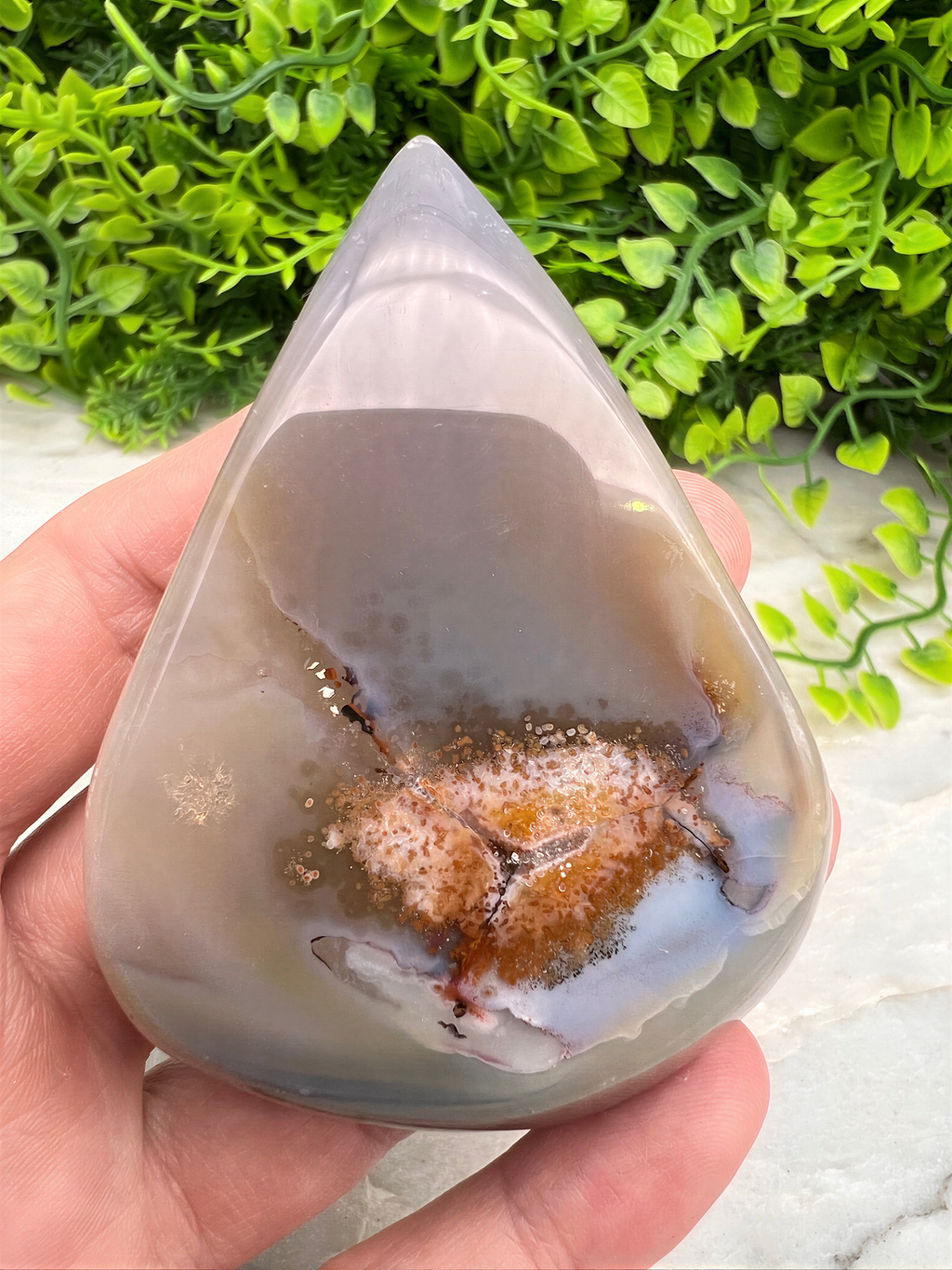 A hand is holding a teardrop-shaped agate stone with a colorful, abstract pattern inside it, set against a backdrop of greenery.