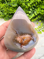 A hand is holding a teardrop-shaped agate stone with a colorful, abstract pattern inside it, set against a backdrop of greenery.