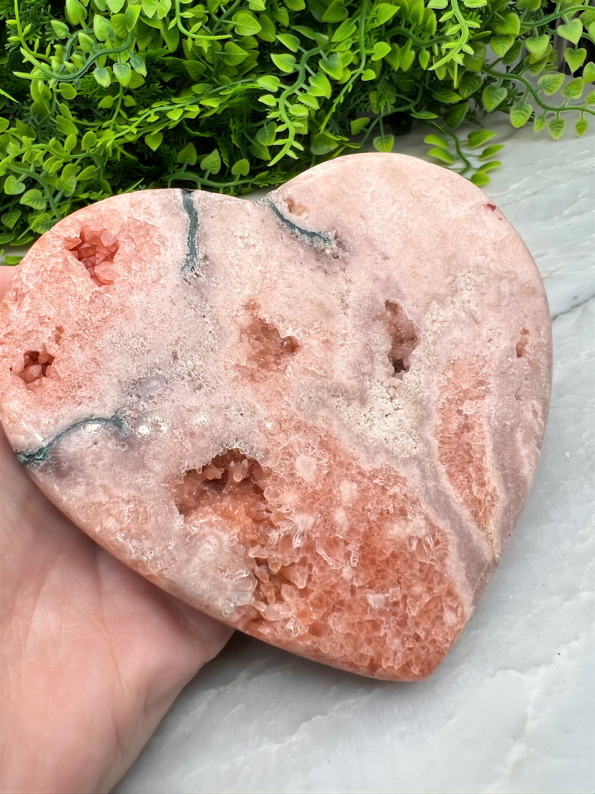 A hand is holding a heart-shaped, pinkish-orange rock with a unique, jagged pattern. The rock is set against a backdrop of green foliage.