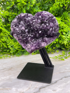 A heart-shaped amethyst crystal is displayed on a black stand, surrounded by greenery.
