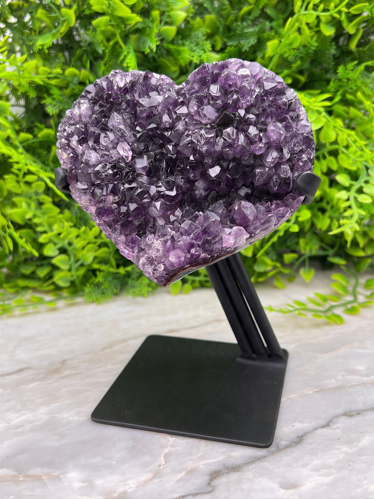 A heart-shaped amethyst crystal is displayed on a black stand, surrounded by greenery.