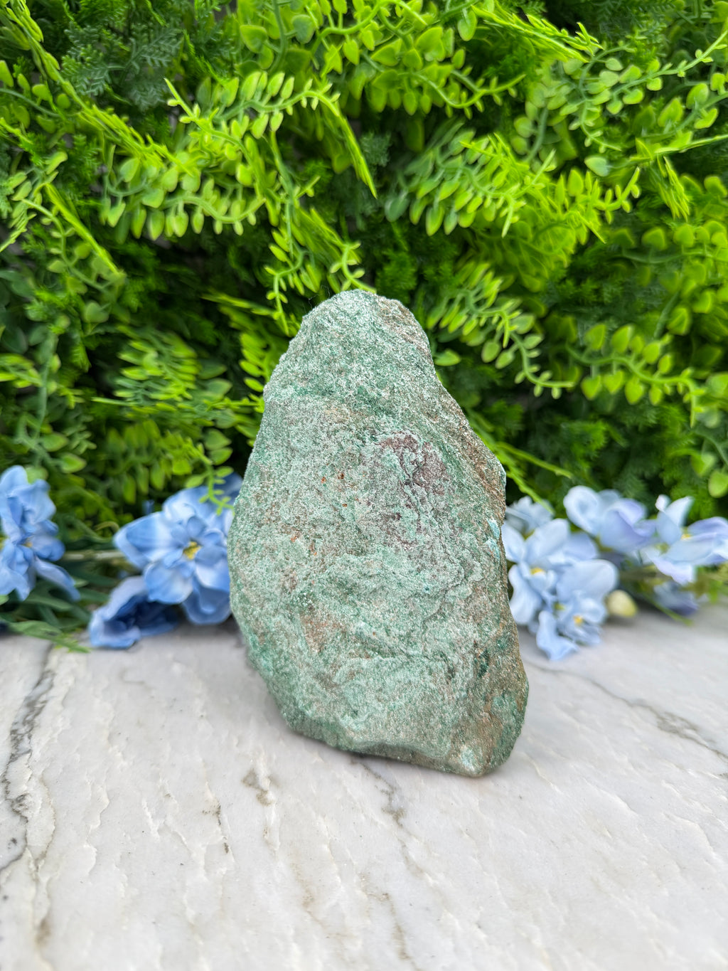 A large, rough-textured rock rests on a bed of green foliage and blue flowers, creating a natural and serene scene.