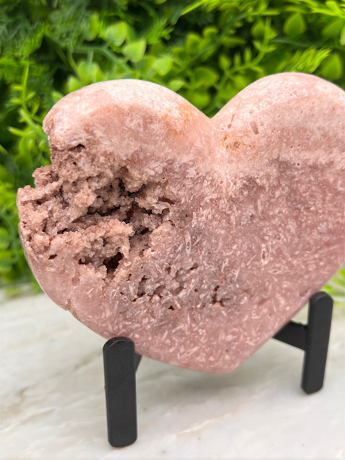 A pink heart-shaped crystal sits on a stand, surrounded by greenery.