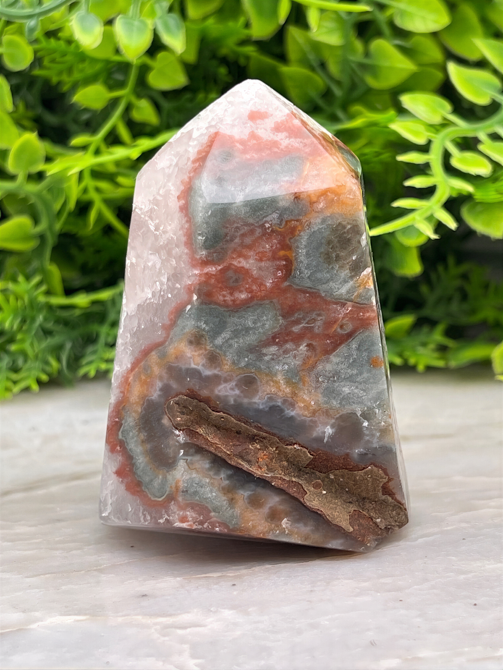 A jagged, multicolored crystal sits on a white surface, surrounded by lush green foliage.