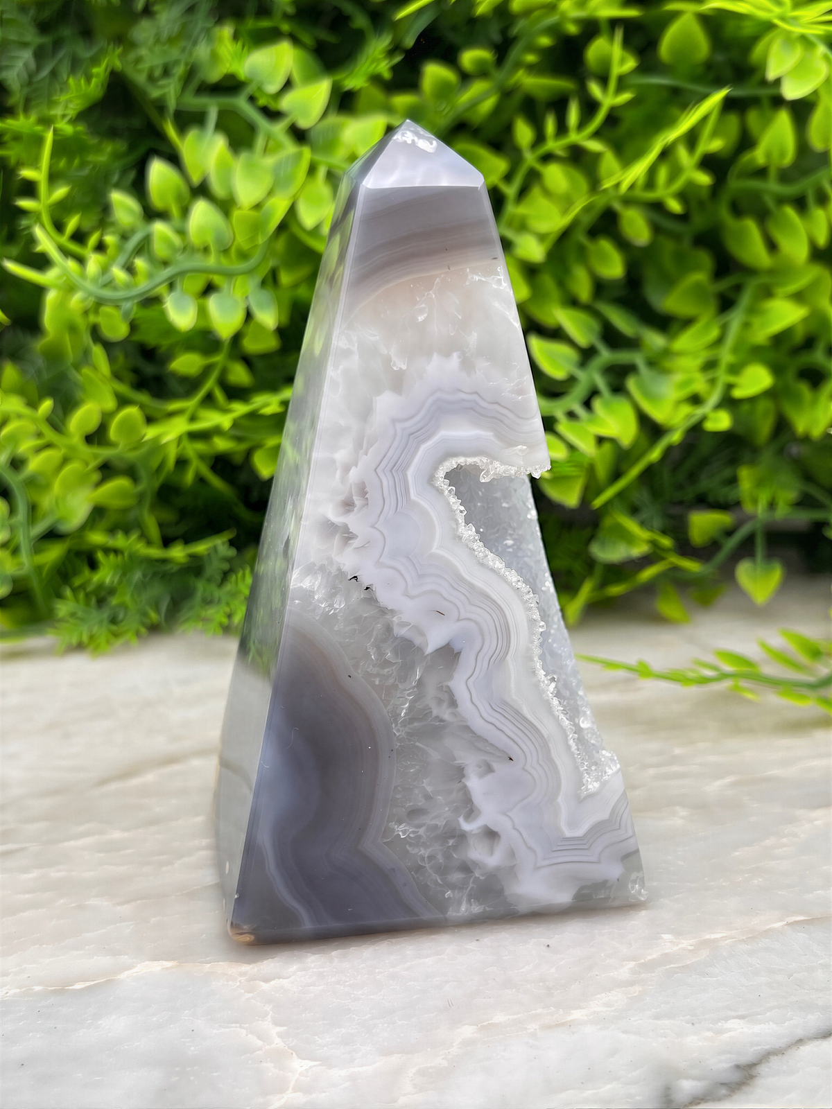 A triangular agate rock formation with a unique, striped pattern rests on a white surface, surrounded by lush green foliage.
