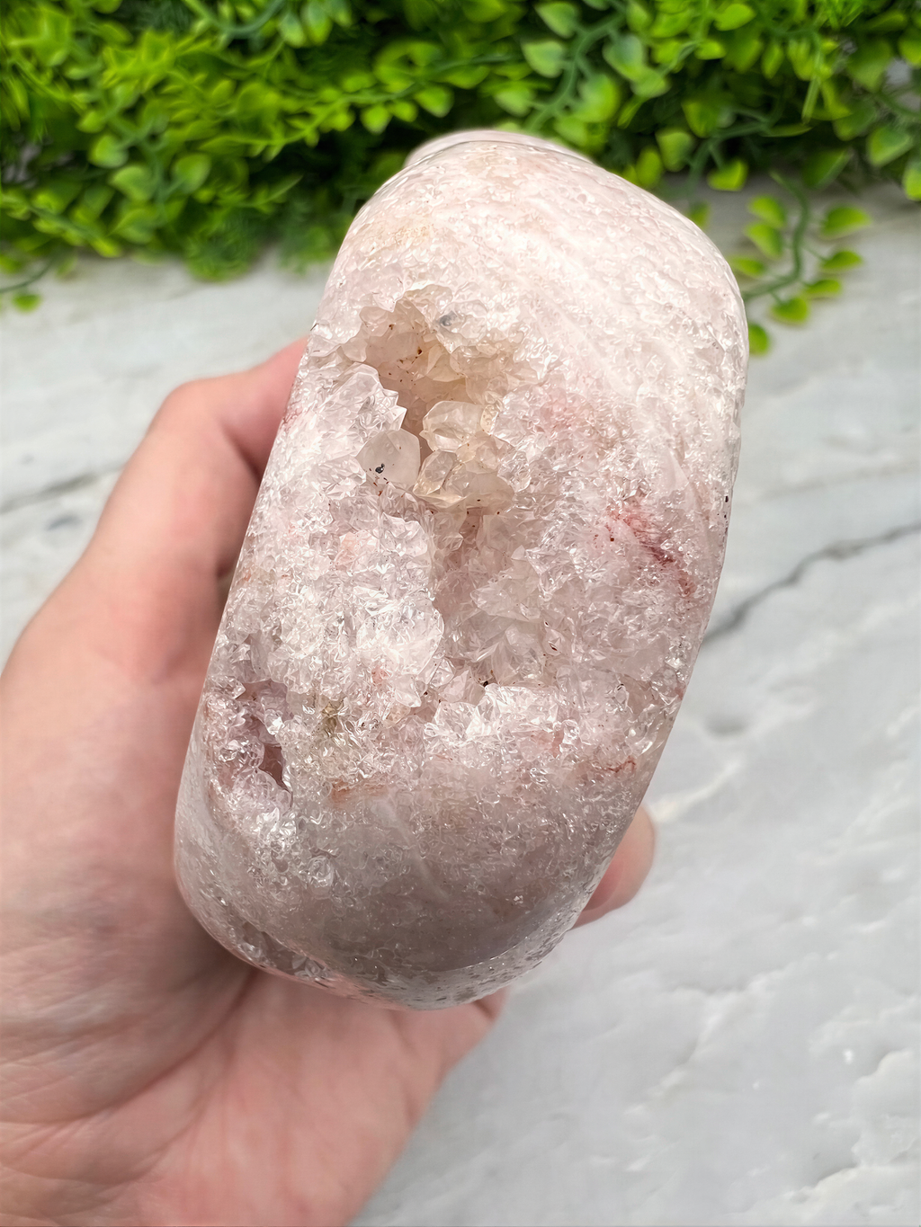 A hand is holding a large, irregularly shaped rock with a pinkish hue, set against a backdrop of greenery.