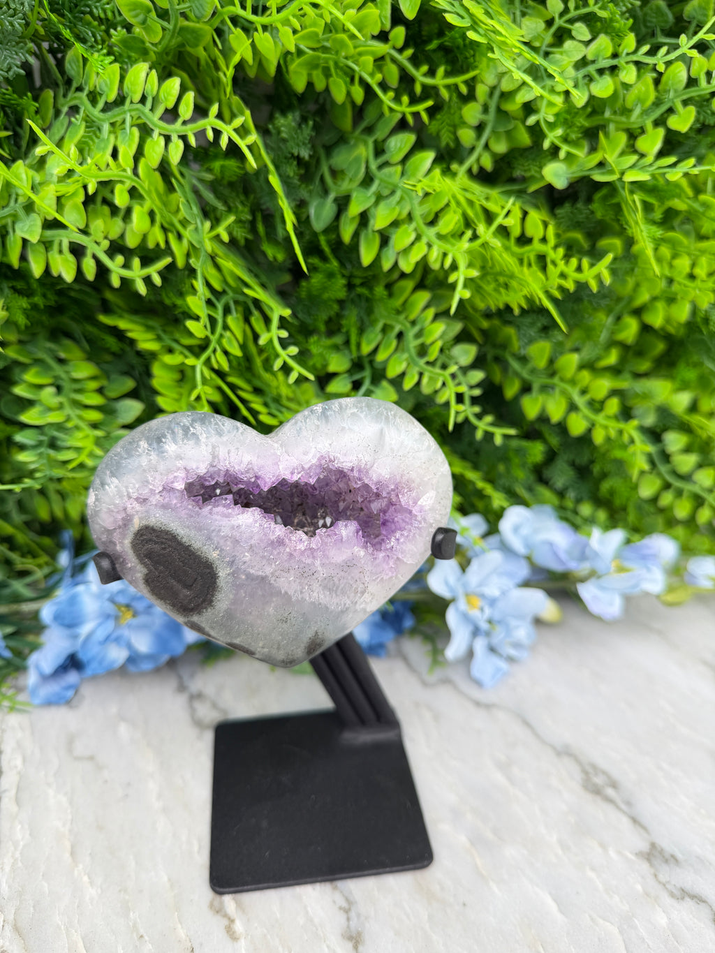 A purple amethyst geode is mounted on a black stand, surrounded by lush green foliage and blue flowers.