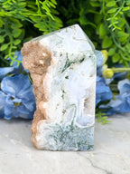 a jade crystal with a unique green and white pattern, placed on a white surface and surrounded by blue flowers.