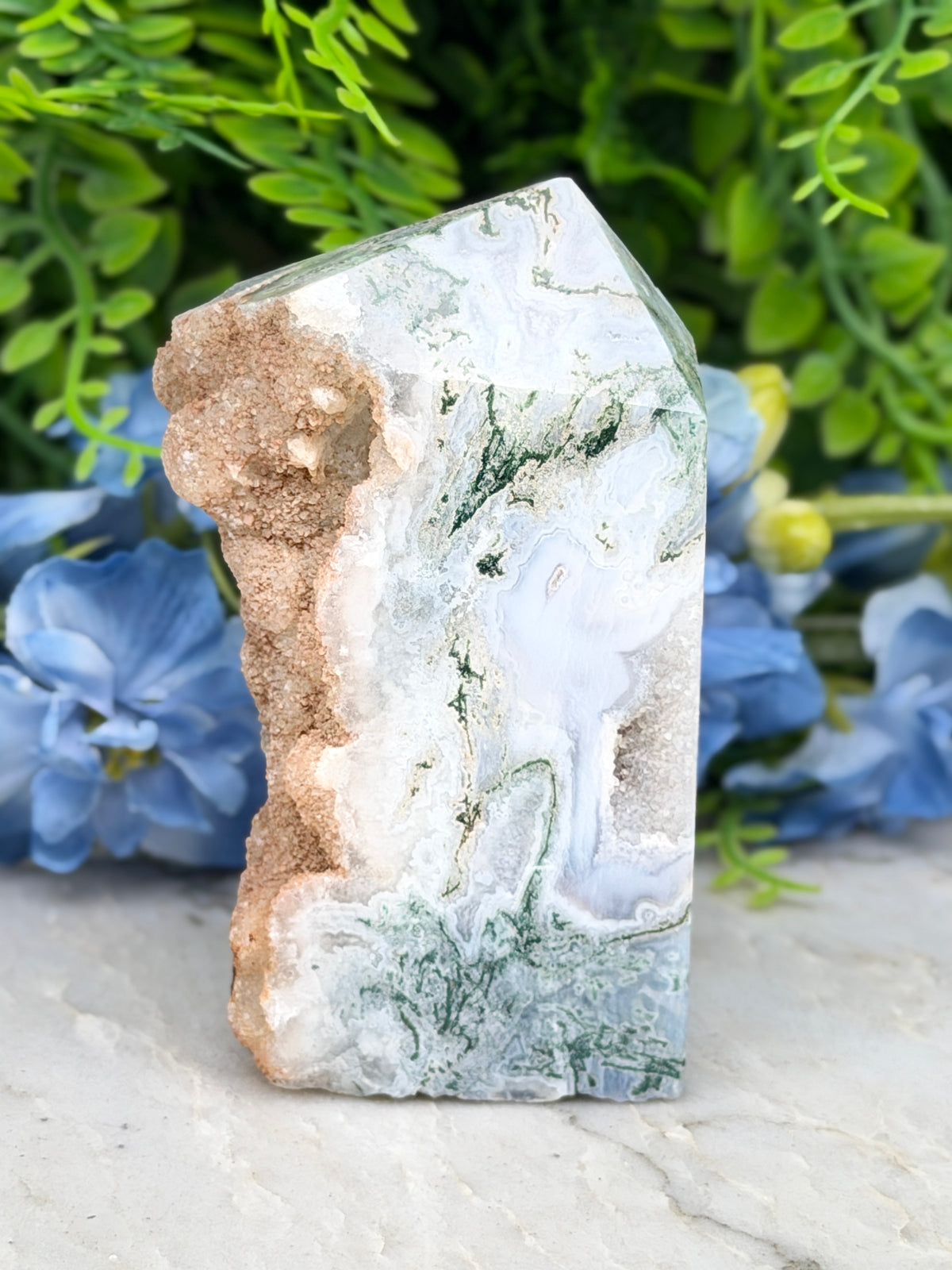 a jade crystal with a unique green and white pattern, placed on a white surface and surrounded by blue flowers.
