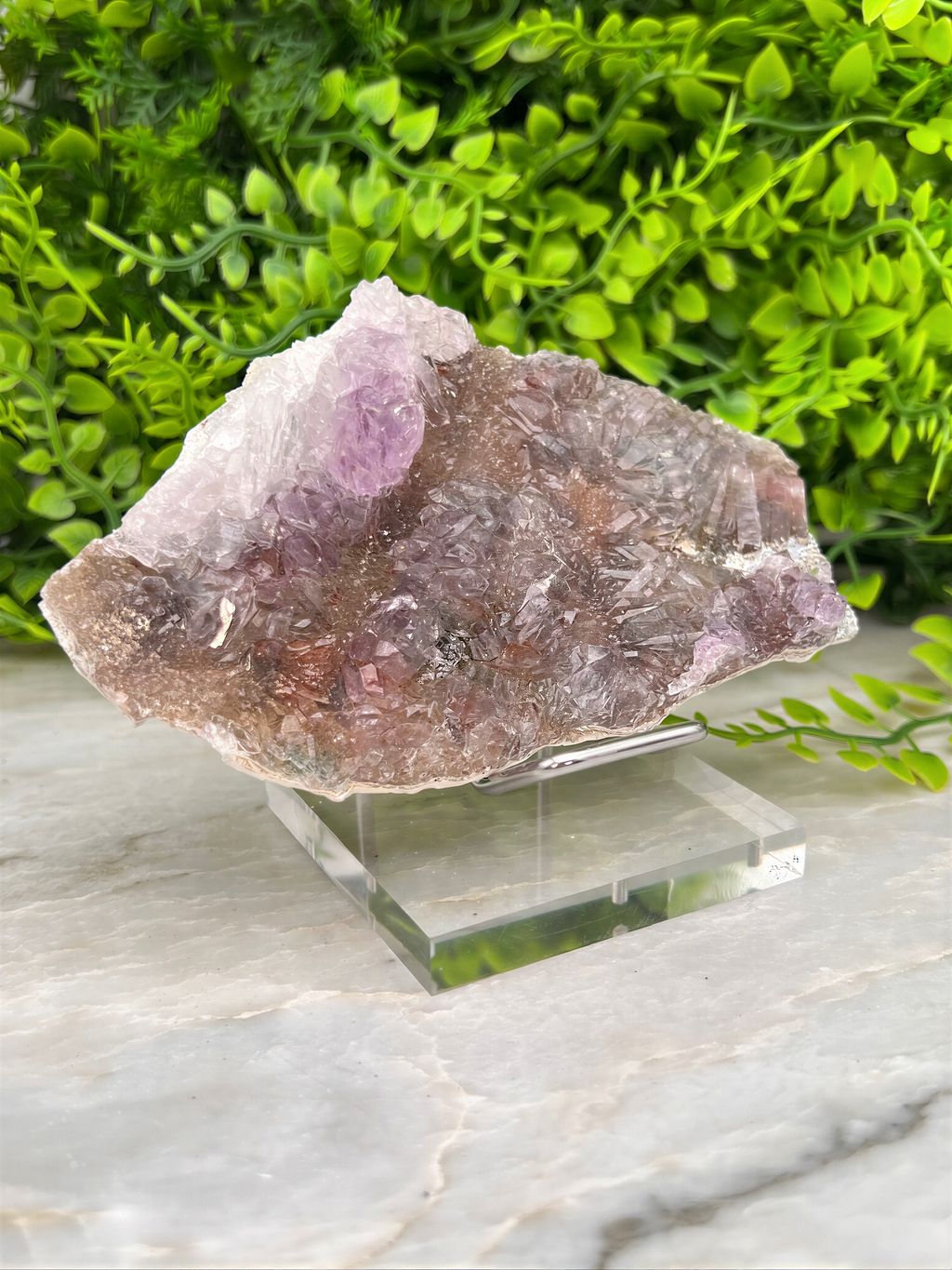 a piece of amethyst crystal placed on a clear glass stand, surrounded by greenery.