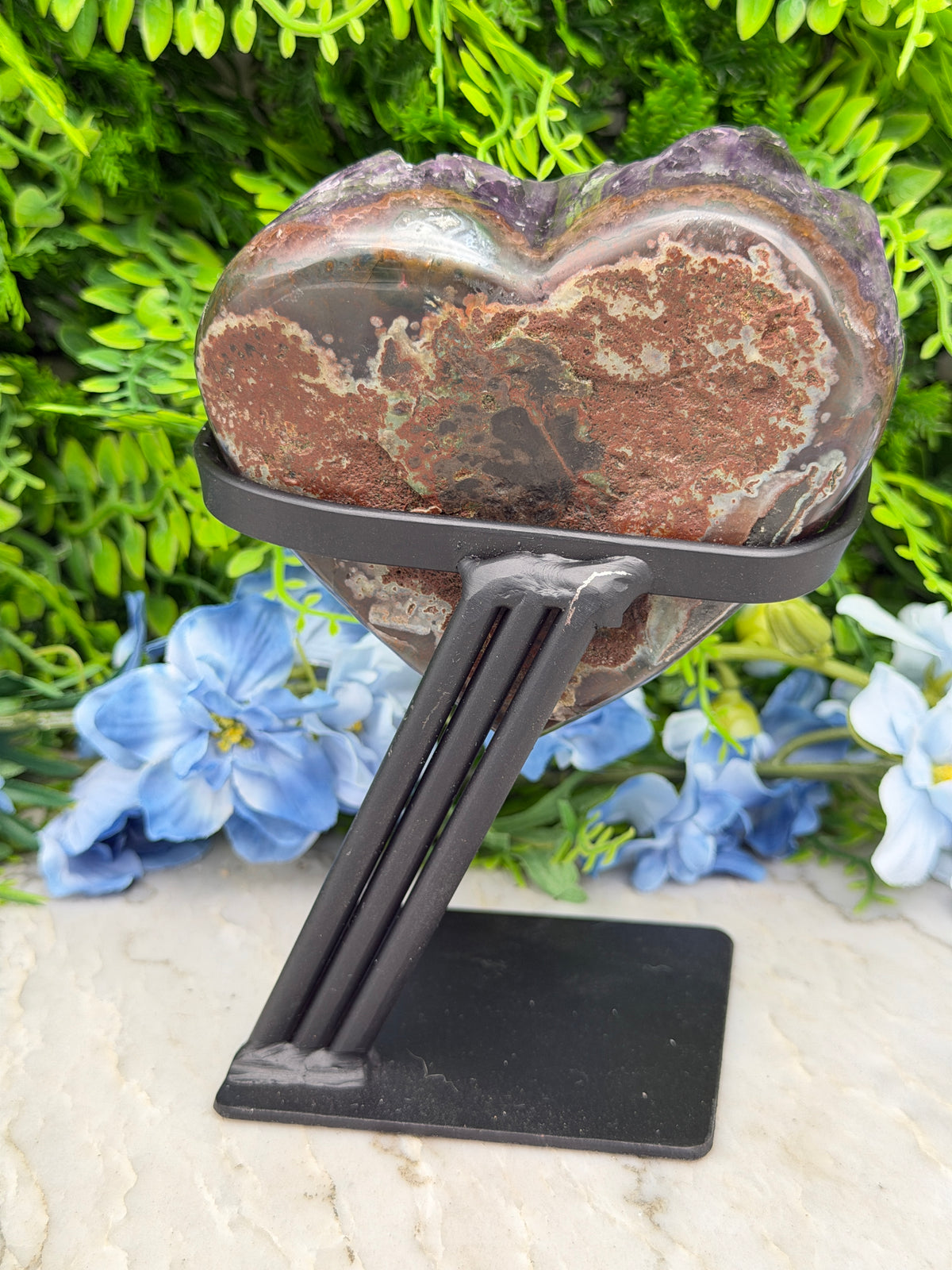 A heart-shaped stone is mounted on a black metal bracket, which is attached to a wall with a black frame. The stone is surrounded by a variety of green plants and blue flowers, creating a natural and vibrant backdrop.