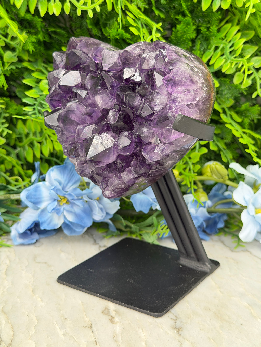 A large purple crystal formation is mounted on a black stand, surrounded by a variety of green plants and blue flowers.