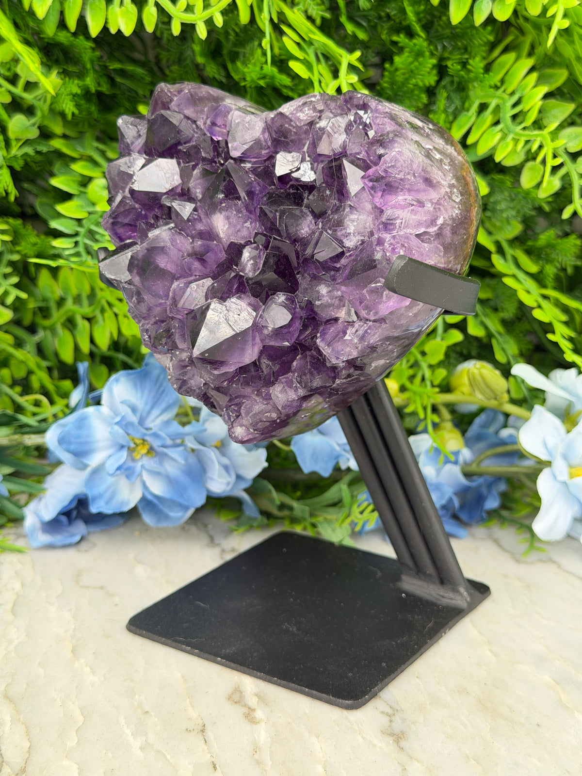 A large purple crystal formation is mounted on a black stand, surrounded by a variety of green plants and blue flowers.