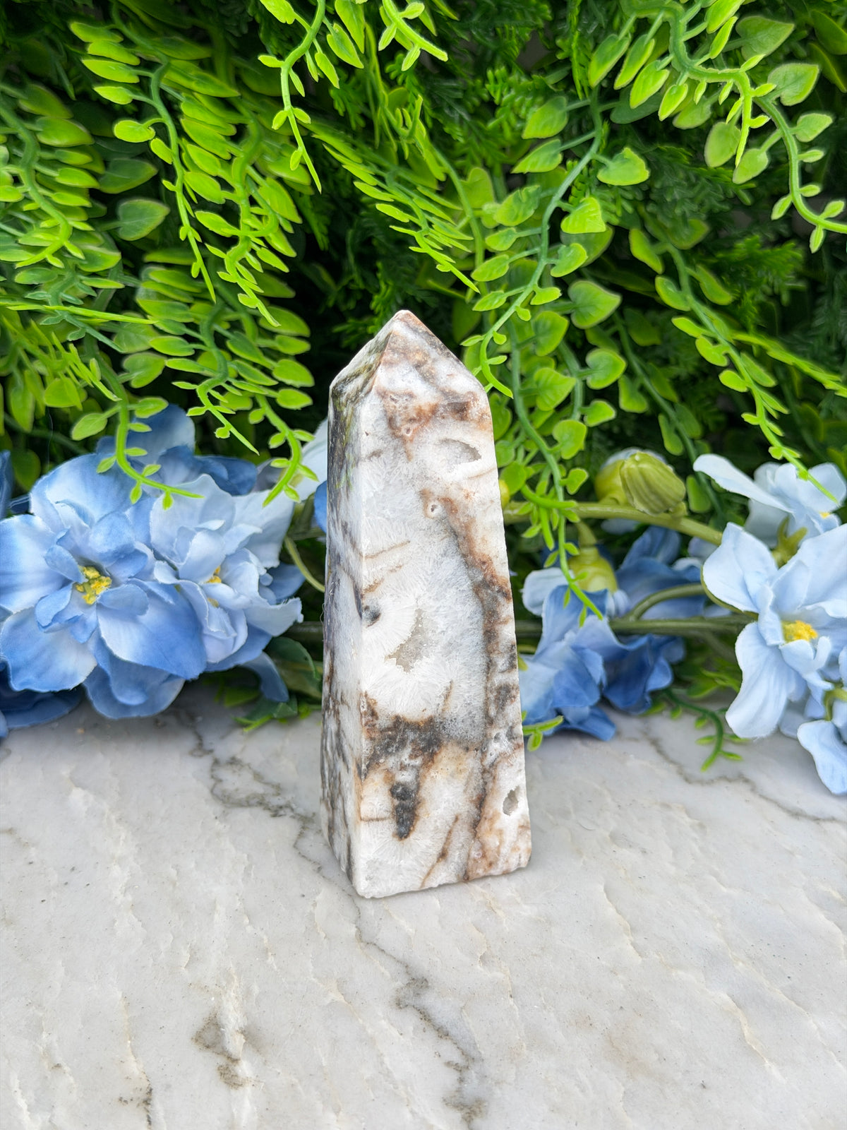 a white stone with a black and brown pattern, surrounded by lush green foliage and blue flowers.