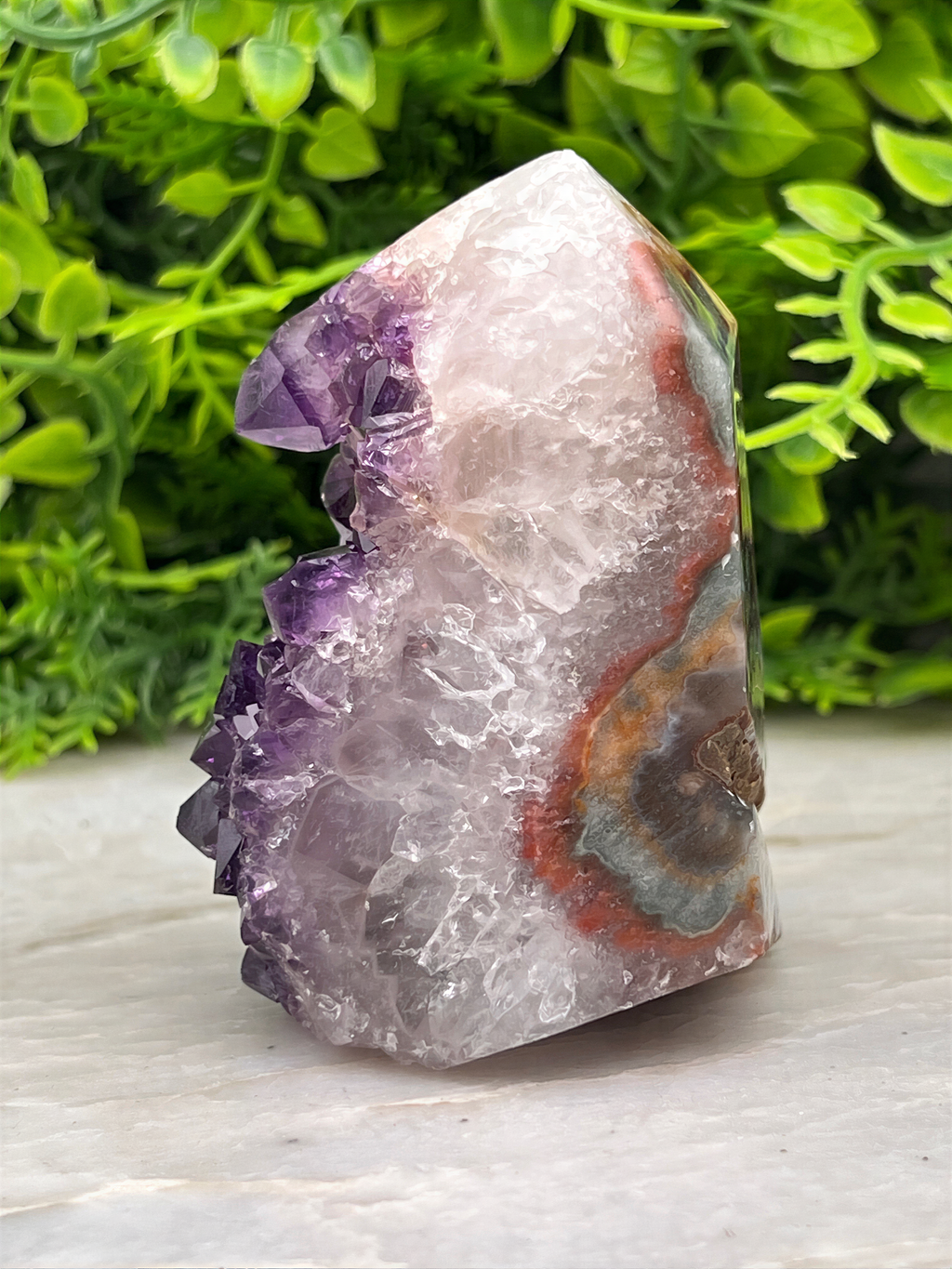 A piece of amethyst, a purple crystal, is displayed on a white surface, surrounded by green foliage.