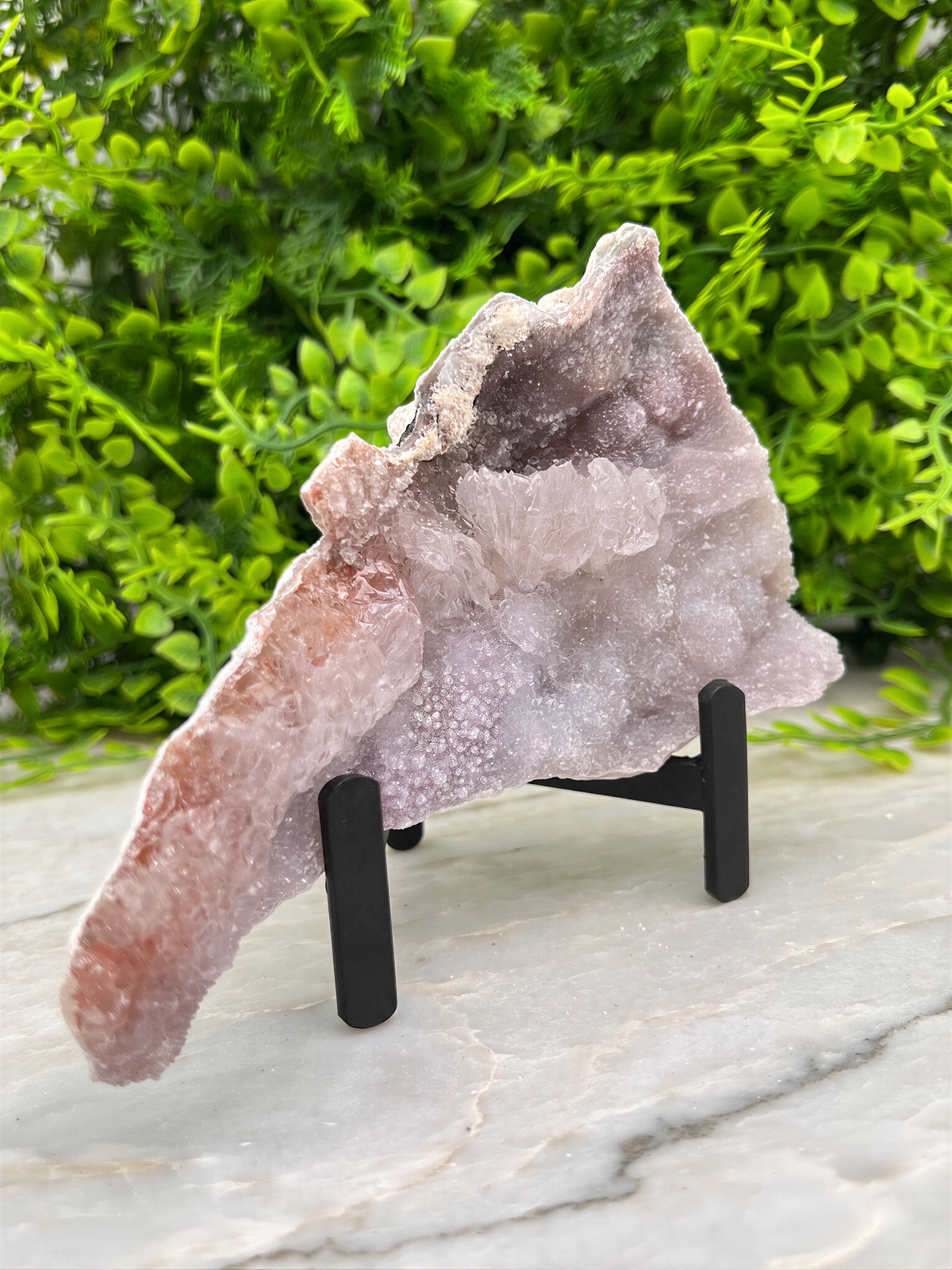 a large piece of amethyst geode placed on a black stand, surrounded by greenery.