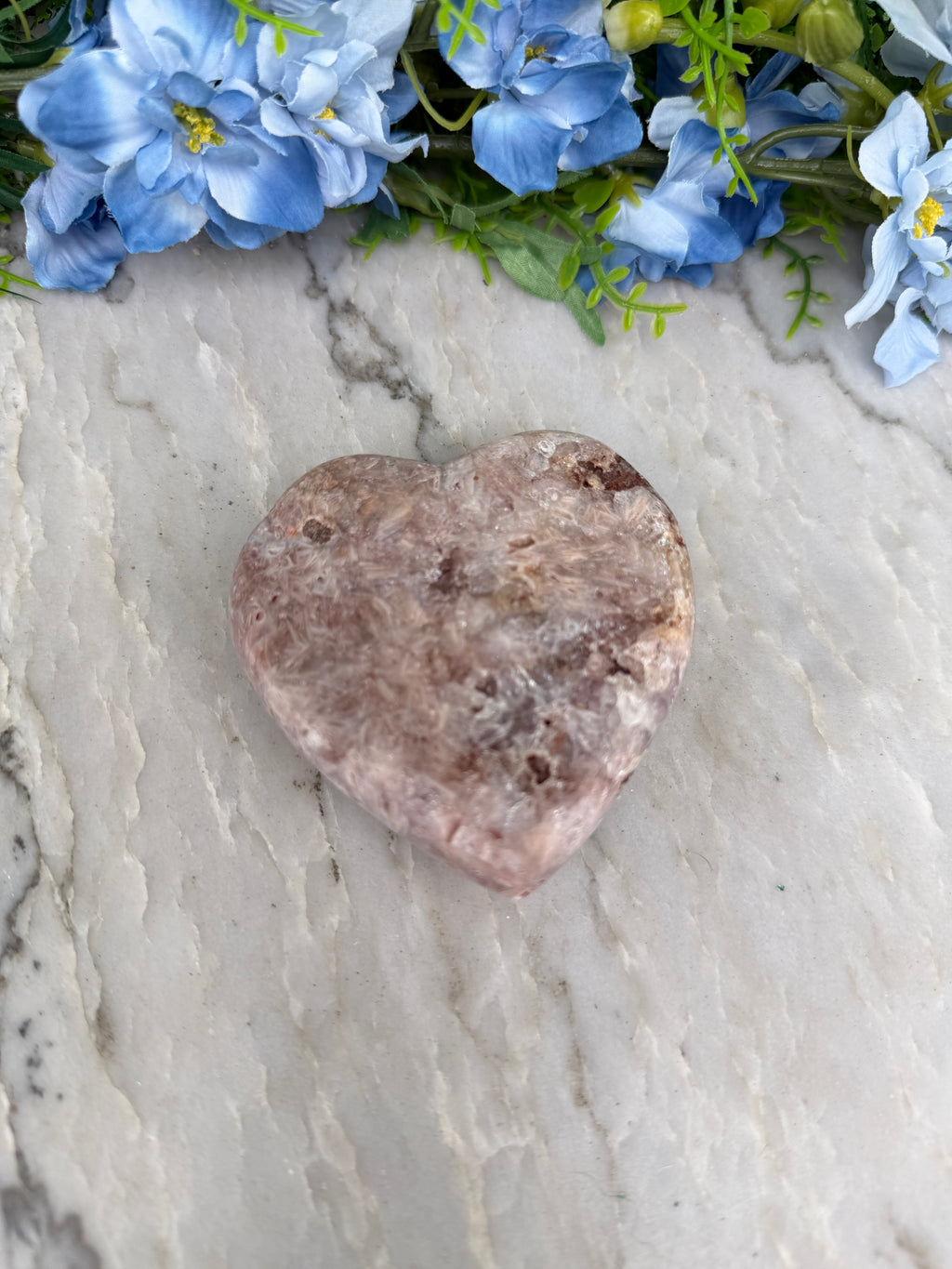 A heart-shaped rock rests on a marble surface, surrounded by blue flowers.