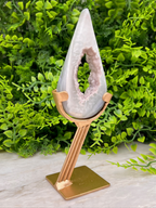 A white geode with a pinkish interior is displayed on a gold stand, surrounded by lush green foliage.