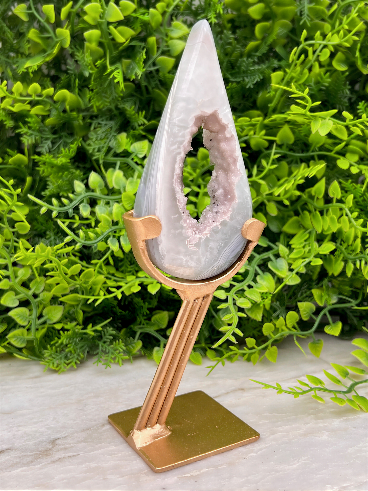 A white geode with a pinkish interior is displayed on a gold stand, surrounded by lush green foliage.
