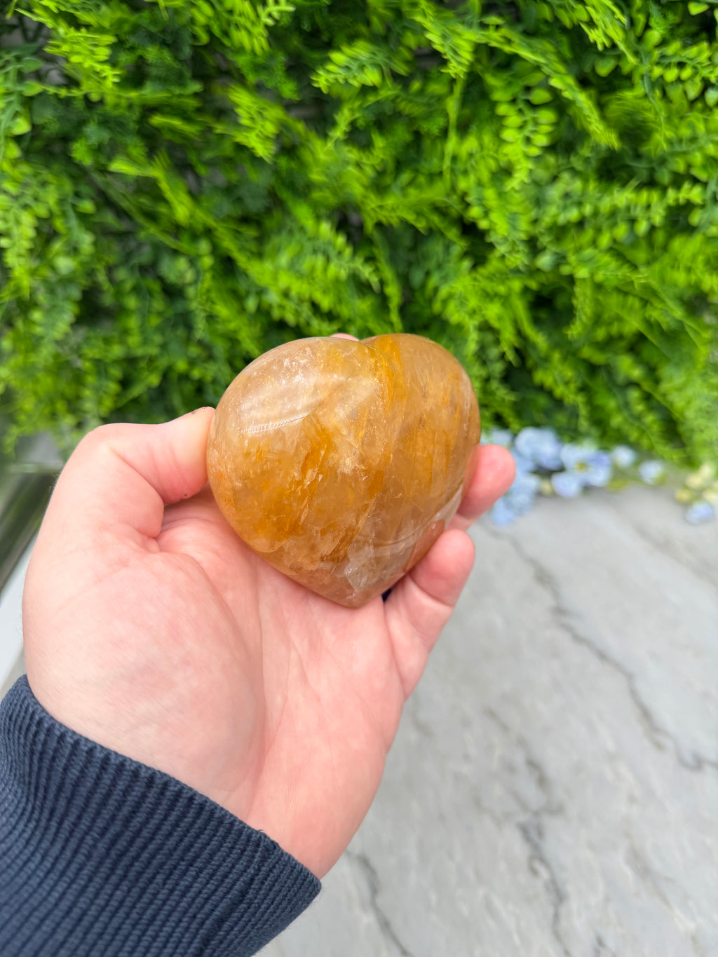 A hand holding a small, round, yellow crystal against a backdrop of green foliage.