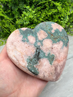 A hand is holding a heart-shaped rock with a green and pink color pattern, set against a backdrop of green foliage.