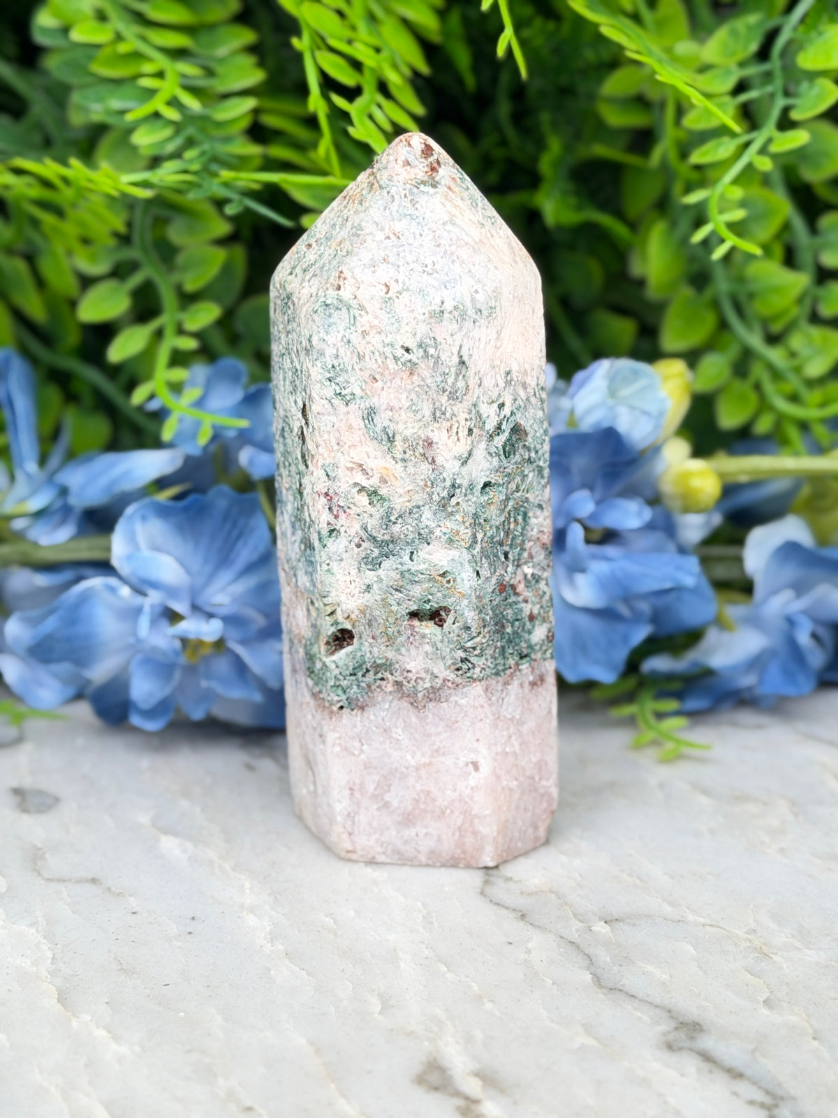 A white and green crystal sits on a marble surface, surrounded by blue flowers.