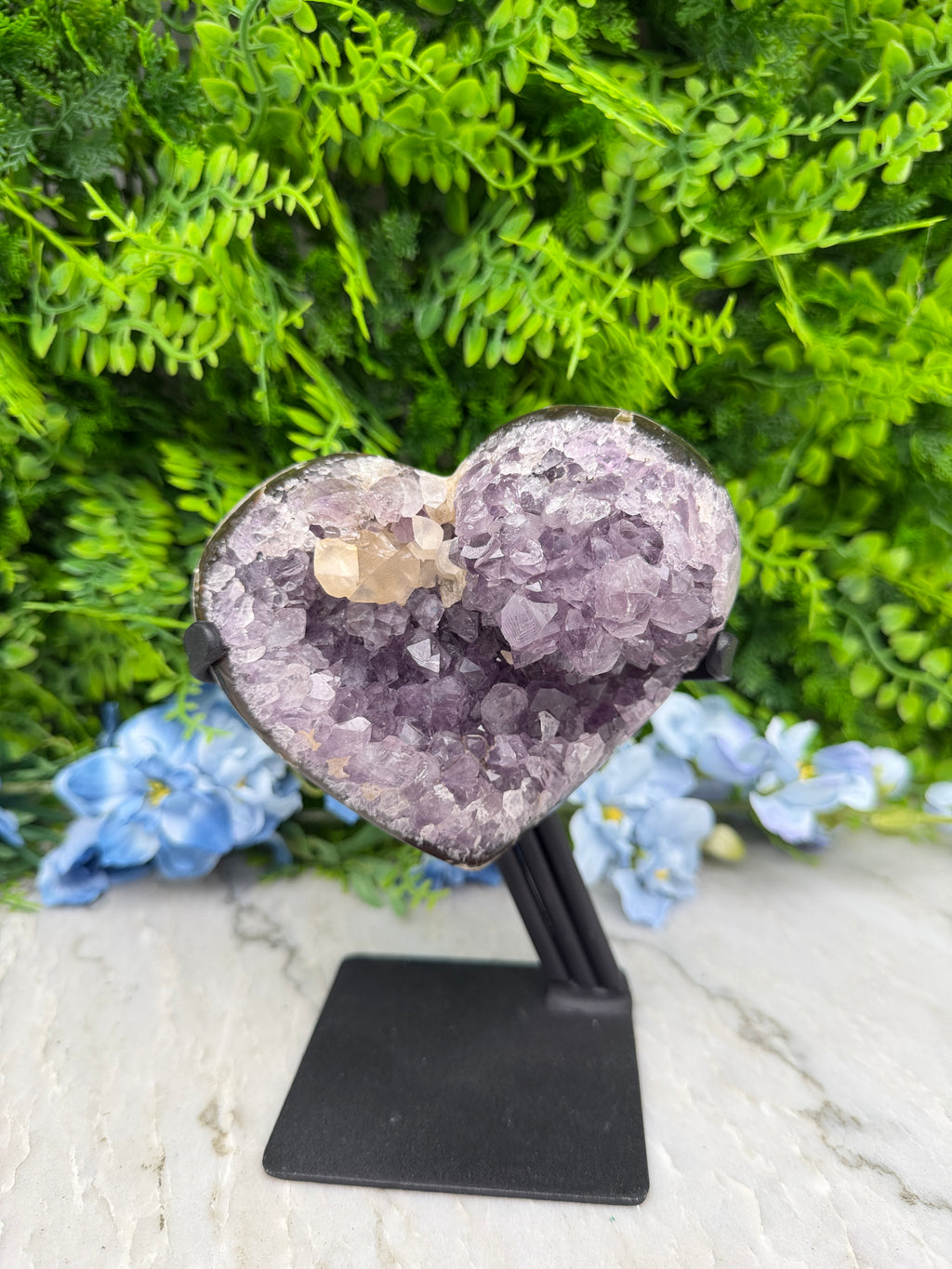 A heart-shaped amethyst crystal is displayed on a black stand, surrounded by lush green foliage and blue flowers.
