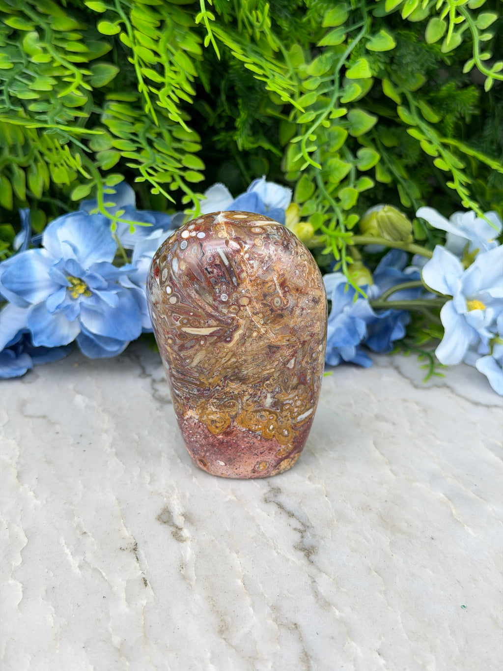 A rock with a unique pattern rests on a white surface, surrounded by lush green foliage and delicate blue flowers.