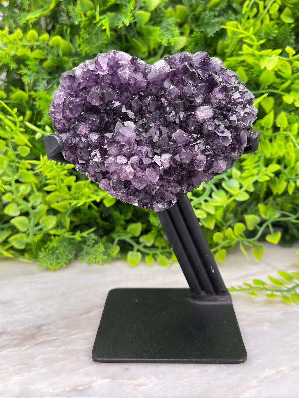 A heart-shaped cluster of purple amethyst crystals is displayed on a black stand, surrounded by lush green foliage.