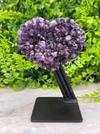 A heart-shaped cluster of purple amethyst crystals is displayed on a black stand, surrounded by lush green foliage.