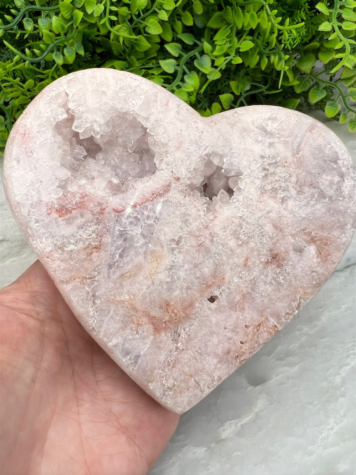 A hand is holding a heart-shaped crystal on a marble surface, with a background of green foliage.