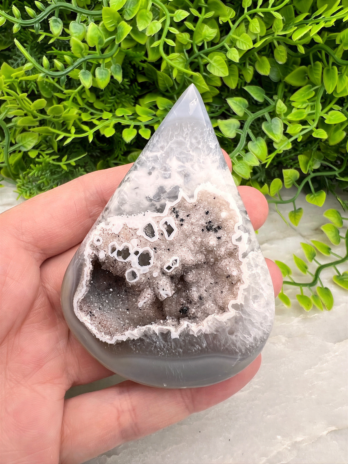 A hand is holding a teardrop-shaped geode with a white, speckled surface, set against a backdrop of green foliage.