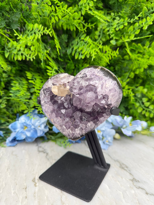 A heart-shaped crystal is mounted on a black stand, surrounded by lush green foliage and blue flowers.
