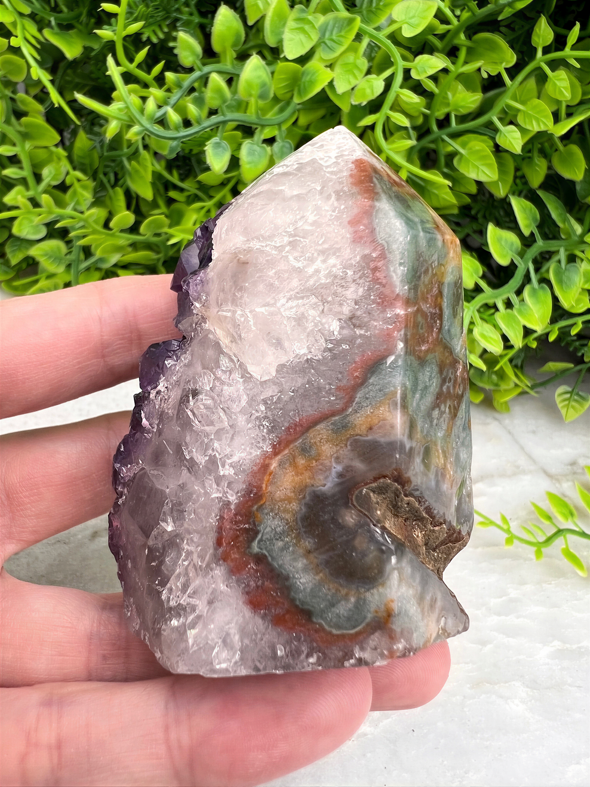 A hand is holding a colorful crystal formation with a mix of white, purple, and green hues, set against a backdrop of lush green foliage.