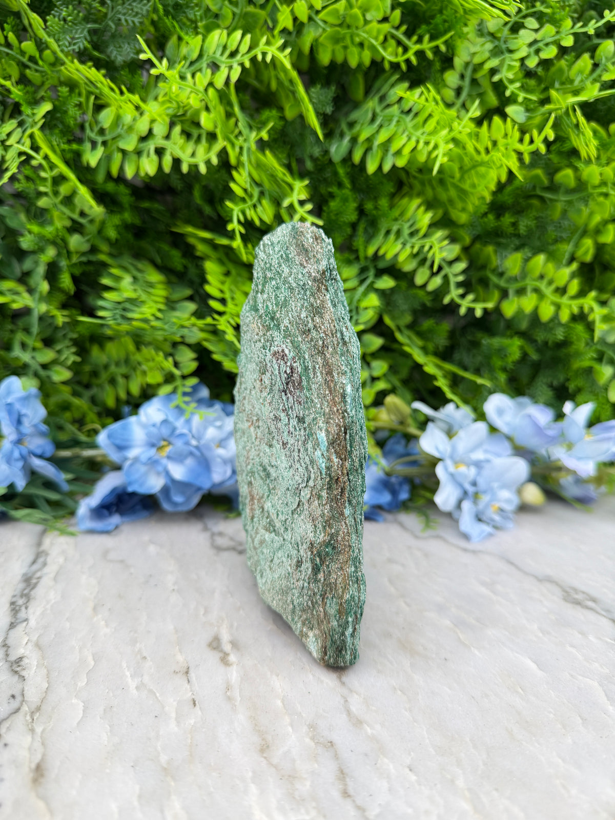 A close-up view of a green rock with a small amount of moss growing on it, set against a backdrop of lush green foliage and delicate blue flowers.