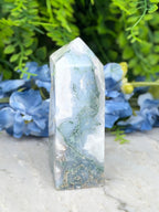 a clear, jagged crystal with a greenish-blue hue, set against a backdrop of lush, green foliage and delicate blue flowers.