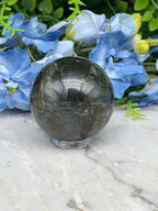 A black gemstone ball rests on a white marble surface, surrounded by vibrant blue flowers.