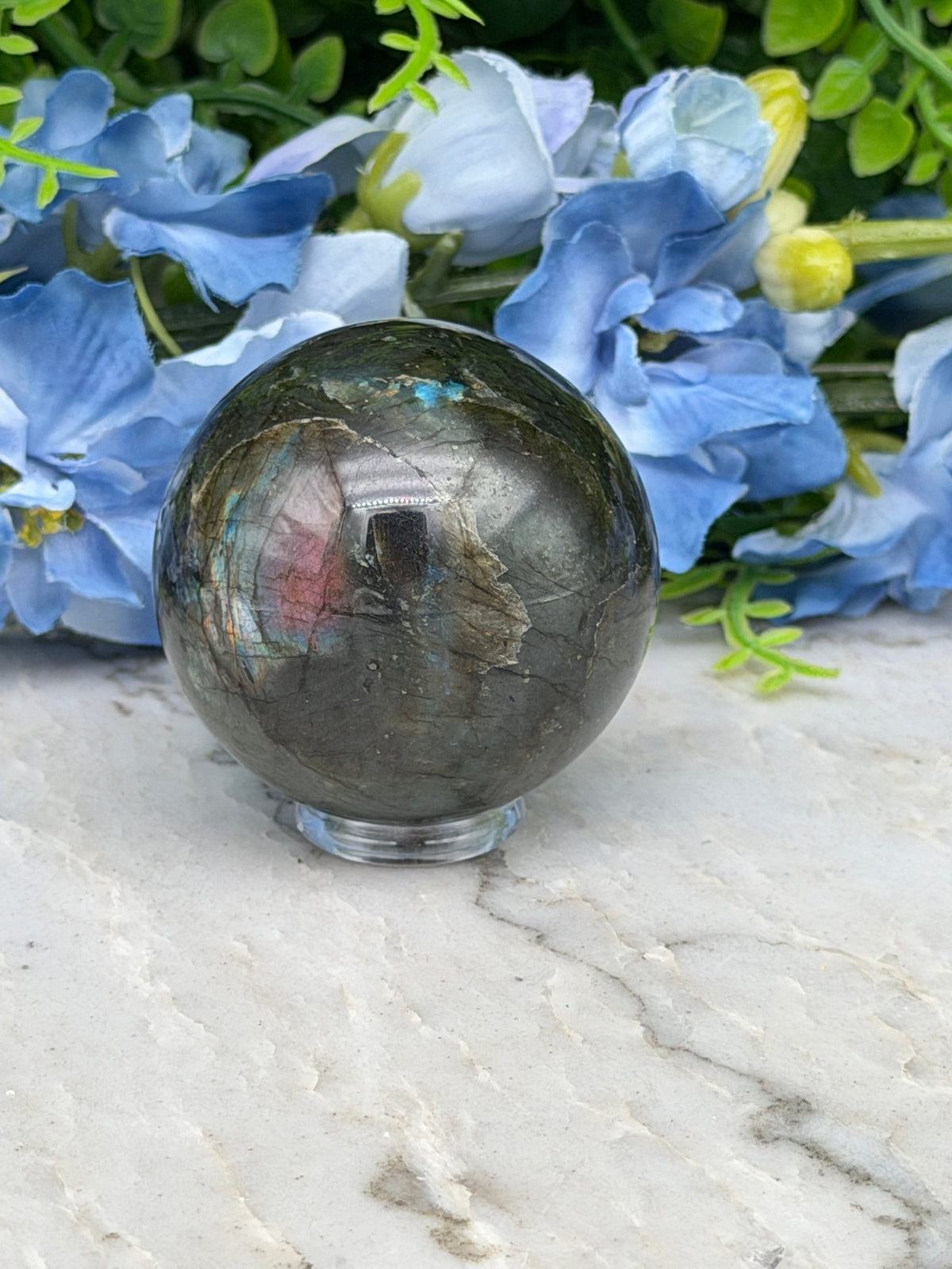 A close-up image of a gray crystal ball surrounded by blue flowers, with a white marble surface in the background.