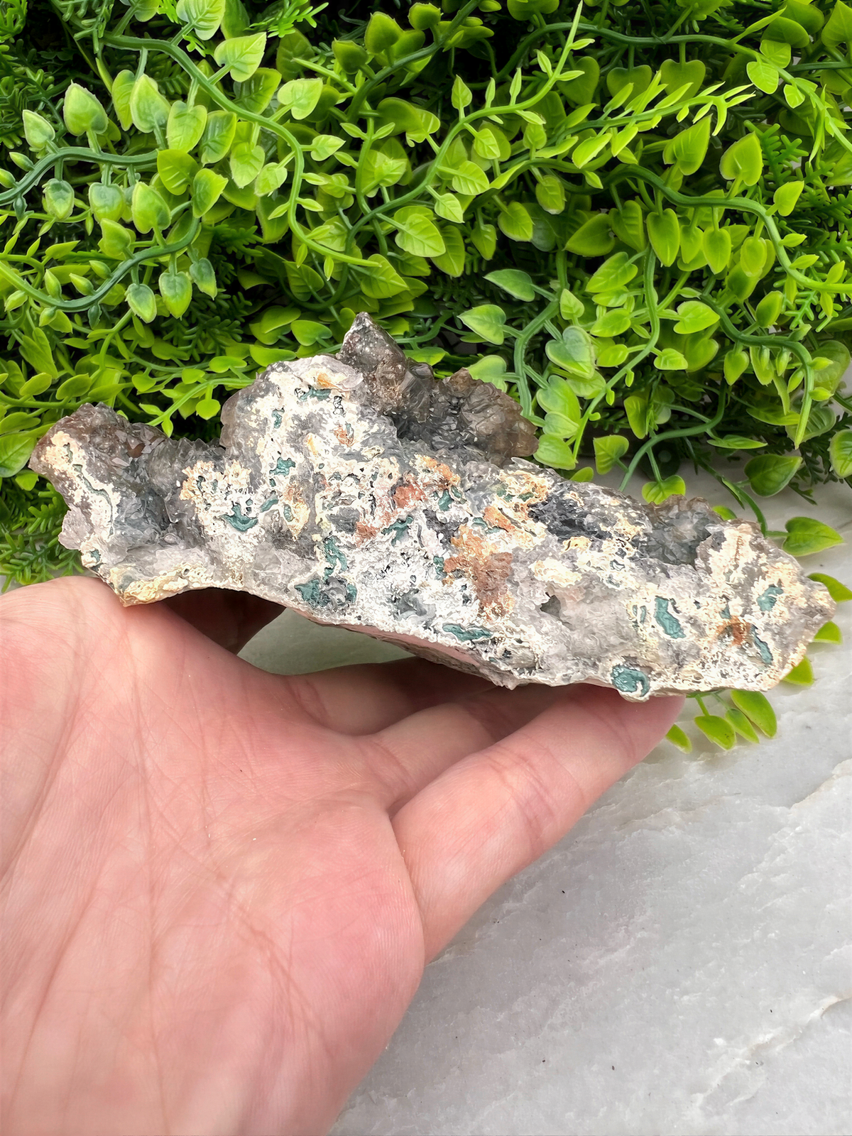 A hand is holding a piece of rock with a greenish hue, set against a backdrop of lush green foliage.
