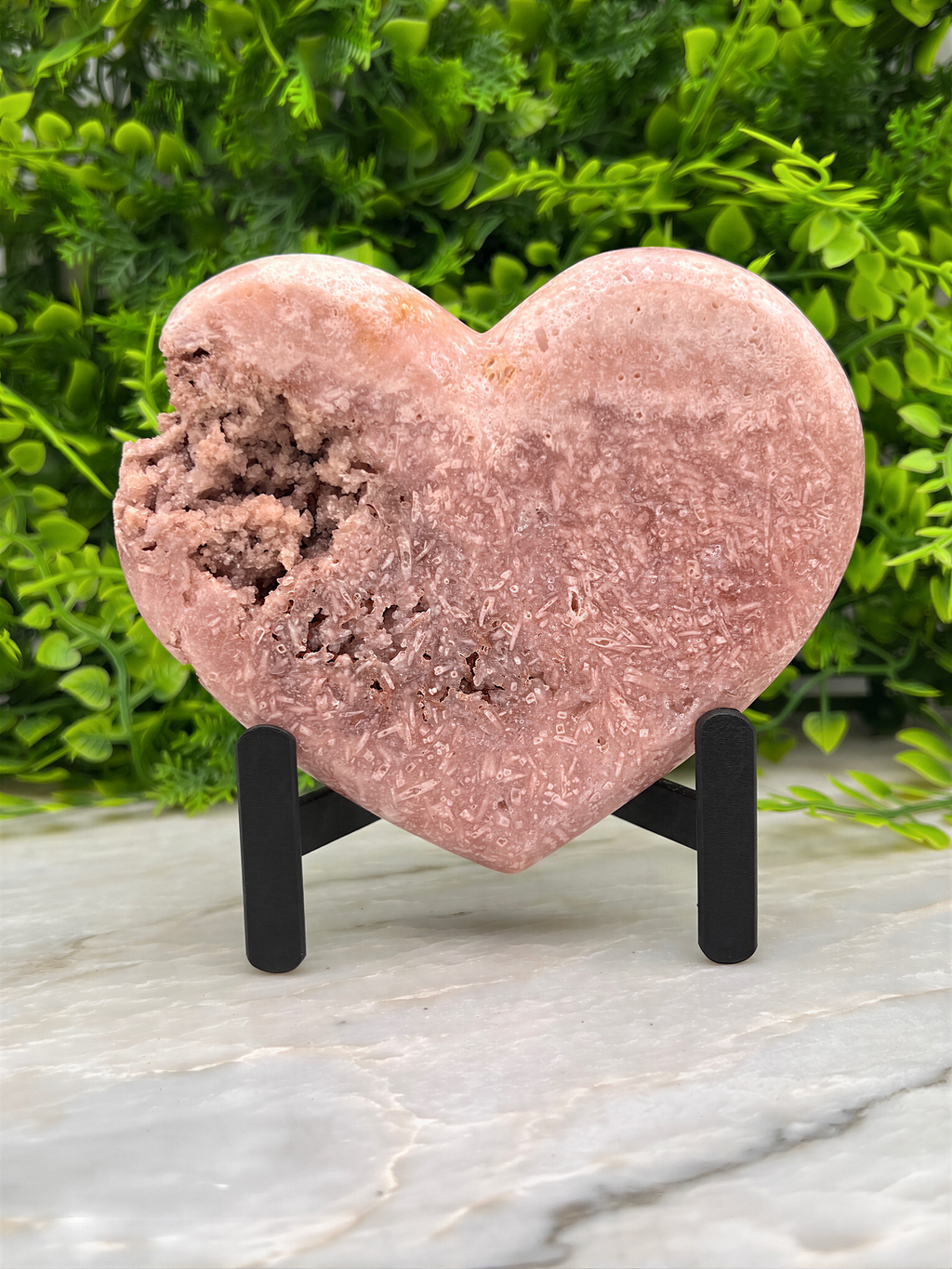 A pink, heart-shaped rock formation is displayed on a stand, surrounded by greenery.
