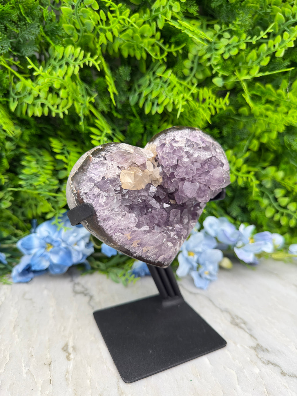 A heart-shaped amethyst crystal is displayed on a black stand, surrounded by lush green foliage and blue flowers.