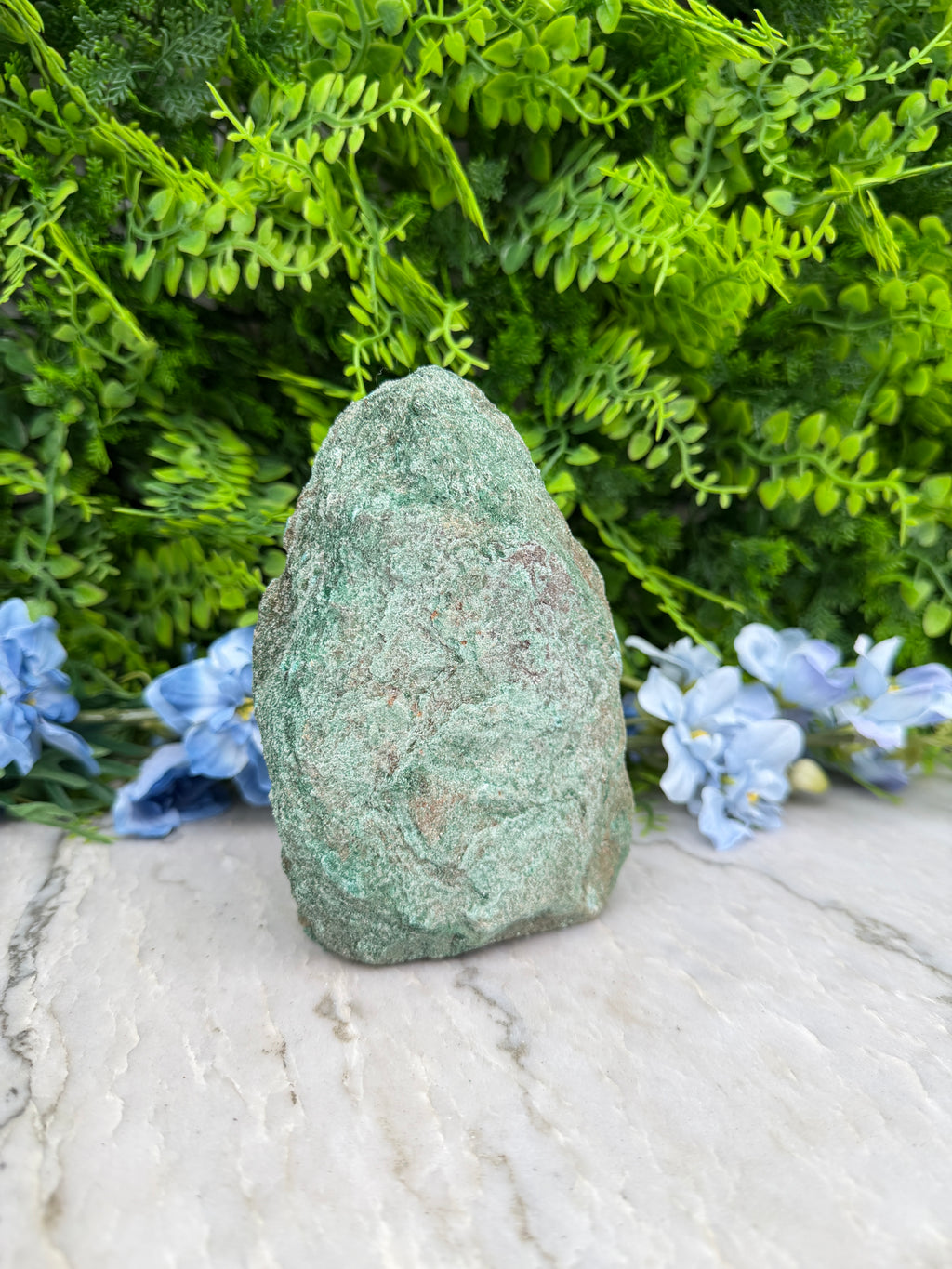 A large, green rock sits in a lush, green garden surrounded by vibrant blue flowers.