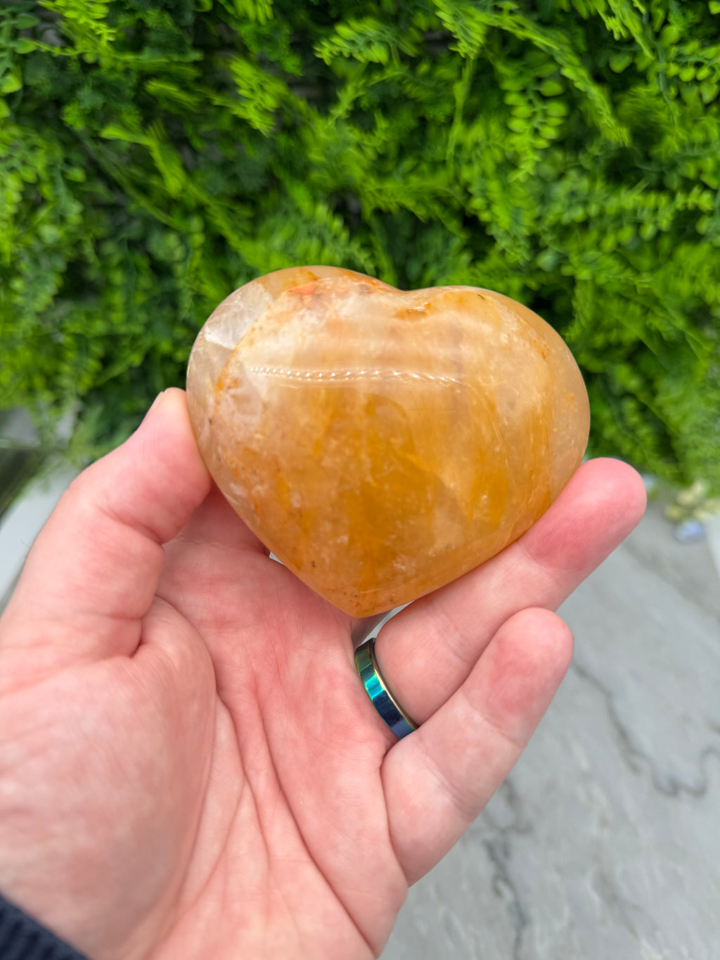 A hand holding a heart-shaped crystal against a backdrop of green foliage.