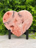 A pink geode-like rock heart-shaped object is displayed on a stand, surrounded by greenery.