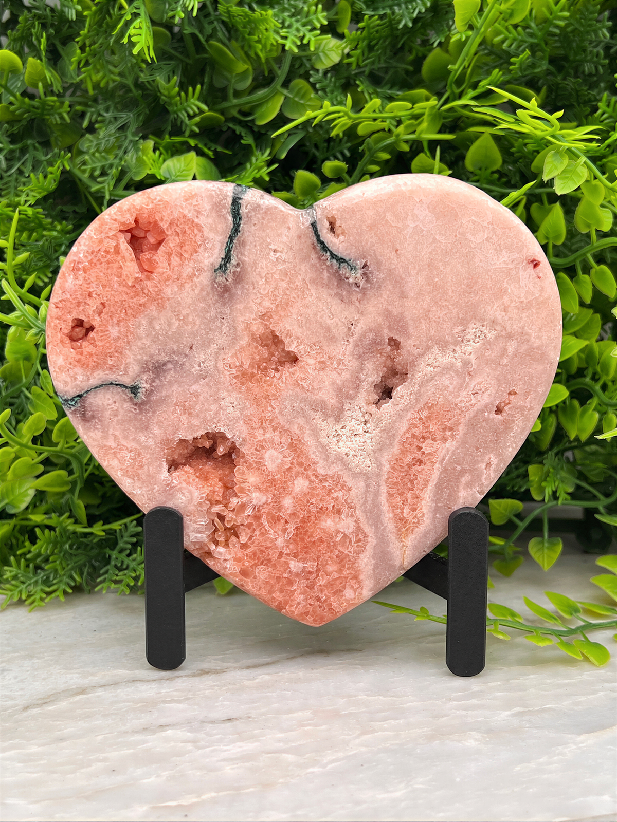 A pink geode-like rock heart-shaped object is displayed on a stand, surrounded by greenery.