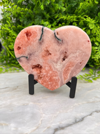 A pink geode-like rock with a heart-shaped pattern is displayed on a stand, surrounded by greenery.