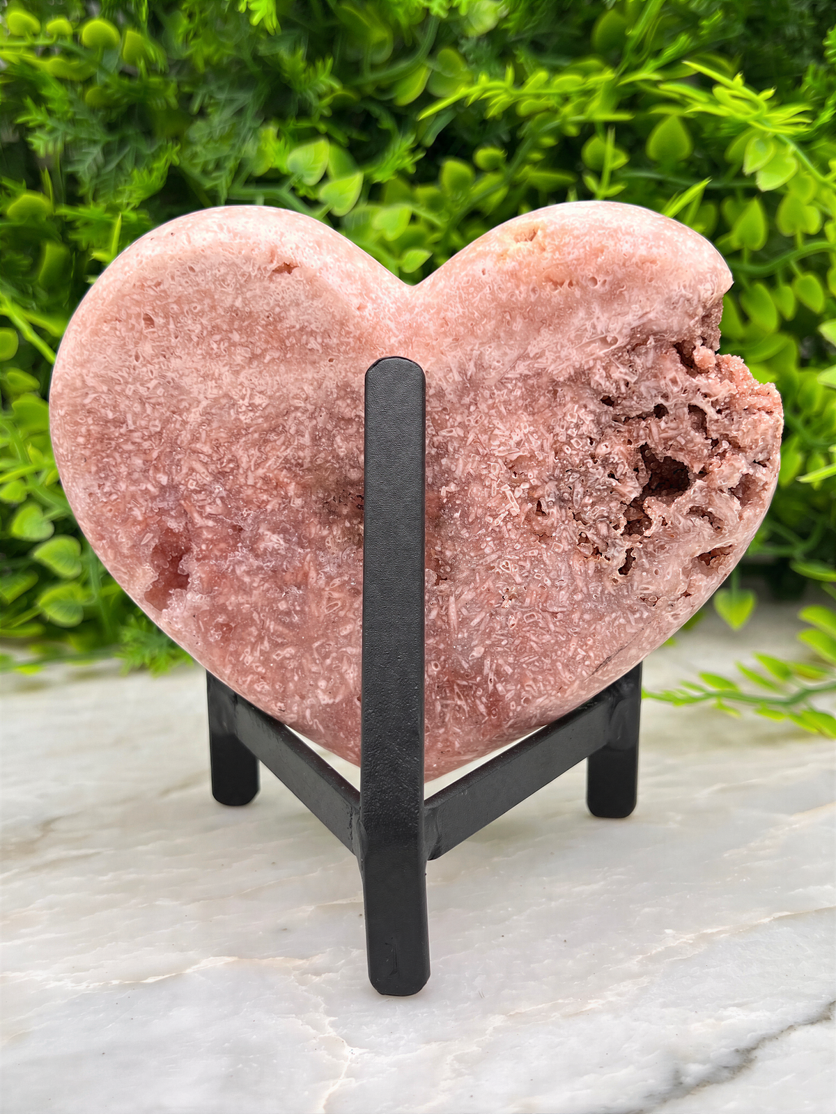 A pink, heart-shaped rock sits on a black stand, surrounded by greenery.
