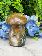 a close-up view of a natural stone mushroom-shaped object, with a striking pattern of orange, brown, and white colors. The mushroom is set against a backdrop of lush green foliage and delicate blue flowers, creating a visually appealing contrast.